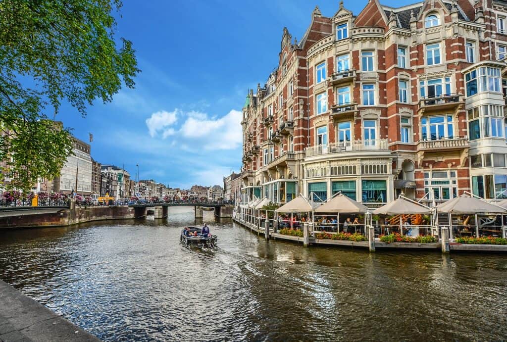 boat going through a canal in Amsterdam with tons of people walking around and old buildings with tons of windows all around