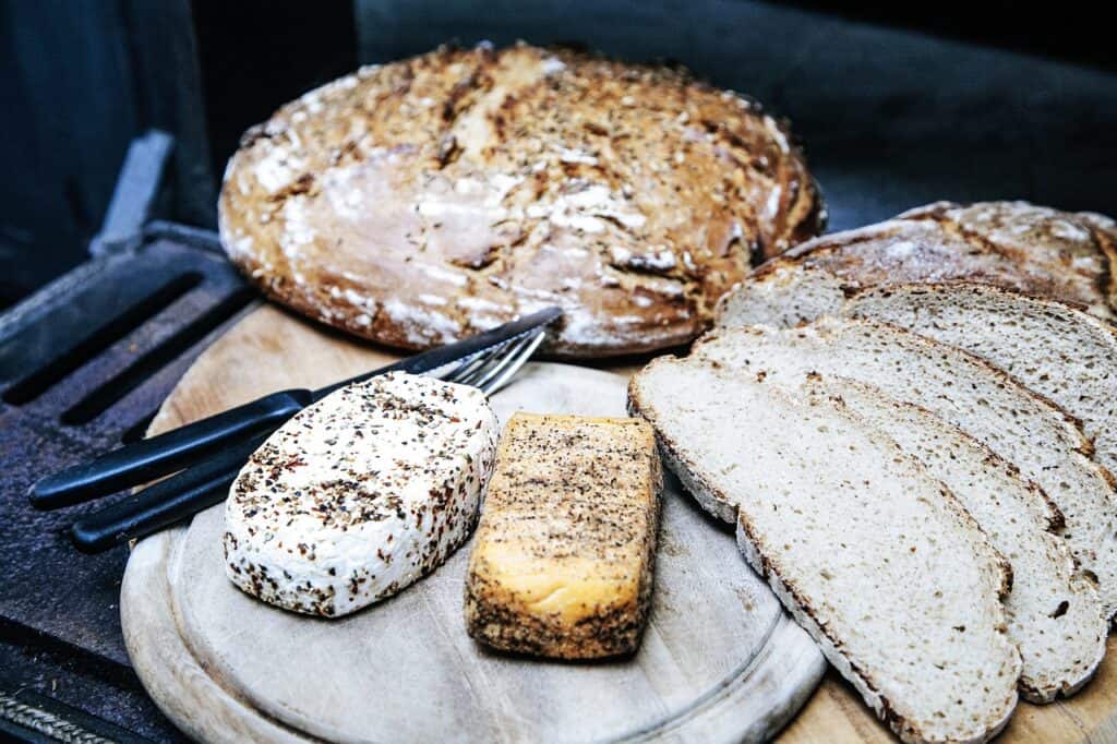 Large loaf and slices of bread next to blocks of cheese on a wooden cutting board