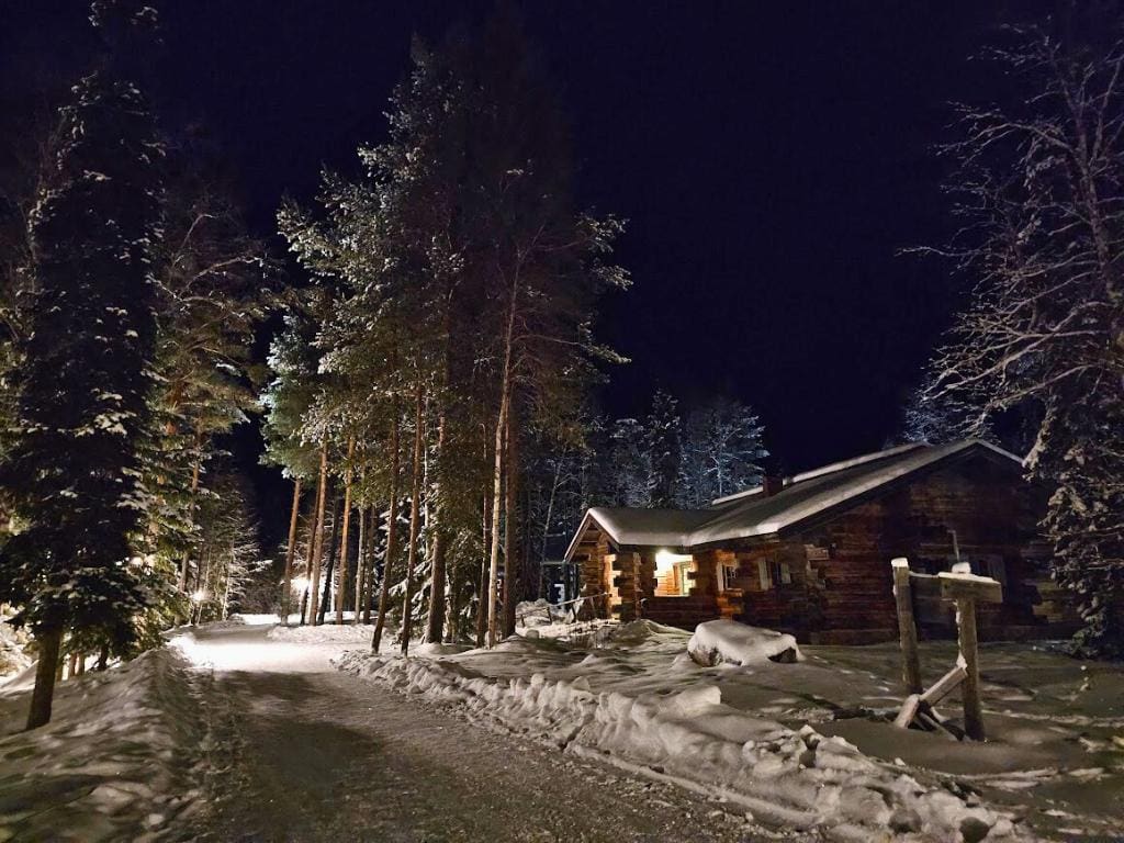 Deep, snowy road surrounded by snowy pine trees and a log cabin at night in Finland